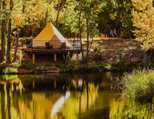 A tent on a deck by a serene pond, surrounded by trees with autumn foliage and two people riding horses nearby.